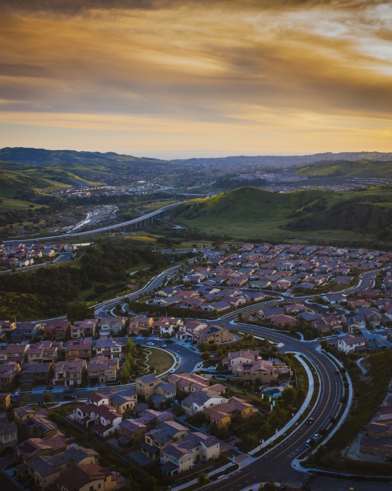 Aerial view of a suburban neighborhood with winding roads, houses, and green hills in the background under a cloudy sky at sunset.